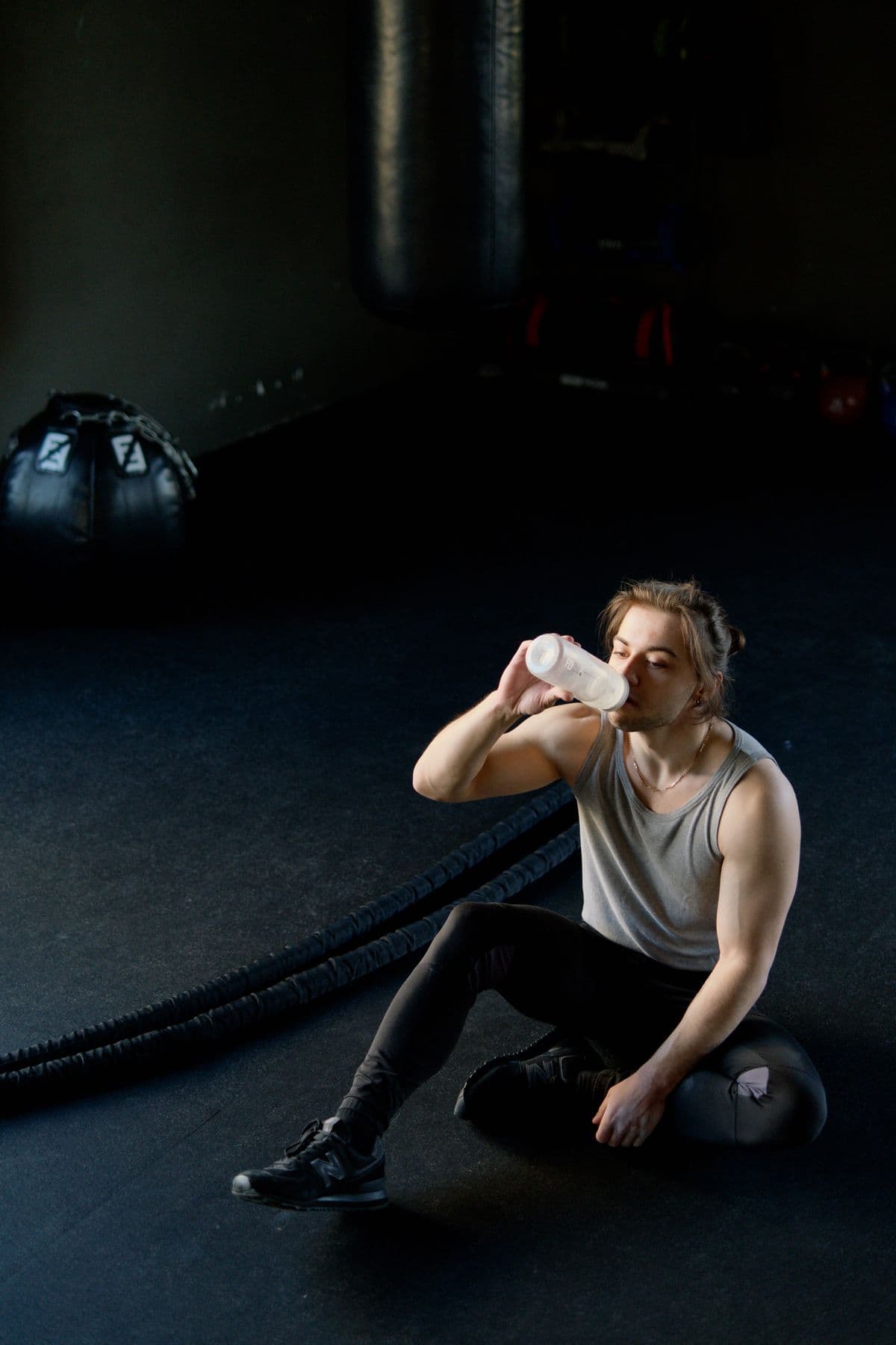 En man i linne sitter på golvet i ett mörkt gym och dricker ur en vit flaska