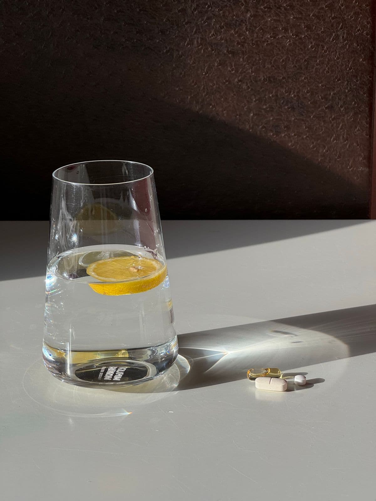 Ett glas vatten med en citronskiva bredvid två vita tabletter och en gul gelékapsel på ett solbelyst vitt bord